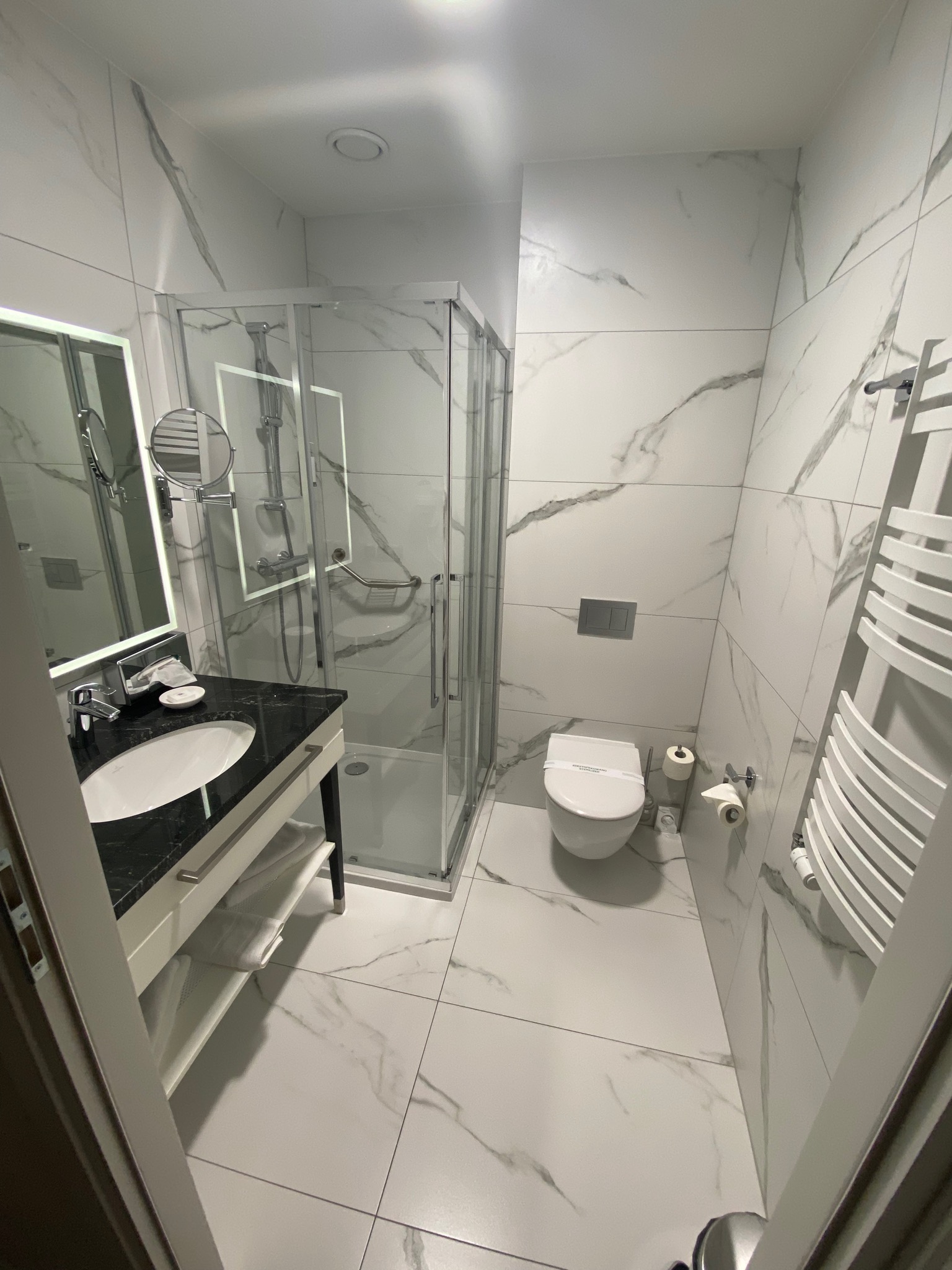 Nowoczesna łazienka z białymi marmurkowymi płytkami, kabiną prysznicową ze szklanymi drzwiami, umywalką z czarnym blatem i podświetlanym lustrem oraz grzejnikiem drabinkowym.