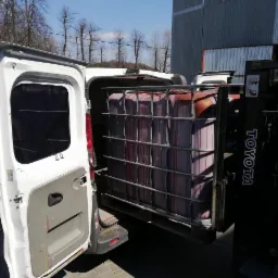 Wózek widłowy Toyota załadowuje kontener IBC z czerwonawą cieczą do białego busa z otwartymi drzwiami, widoczne drzewa w tle.