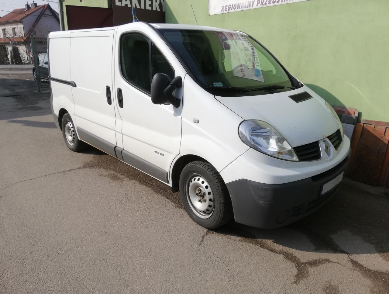Biały samochód dostawczy Renault Trafic zaparkowany na asfalcie przed budynkiem z zieloną ścianą. Na przedniej szybie widoczne ulotki. Widoczne ślady wody na asfalcie.