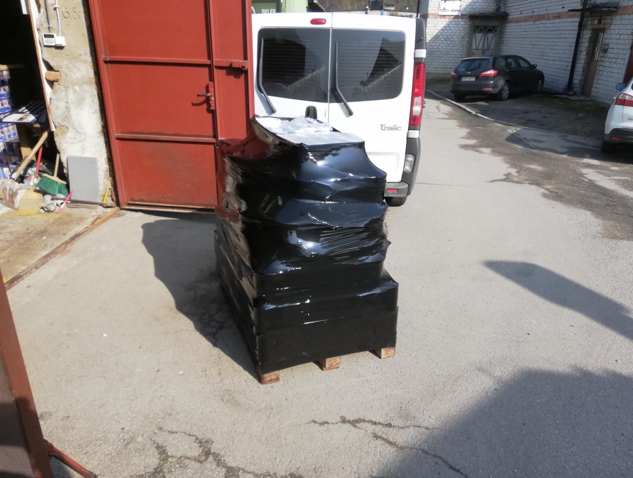 Paczka owinięta czarną folią na palecie, umieszczona za białym busem na zewnątrz budynku.