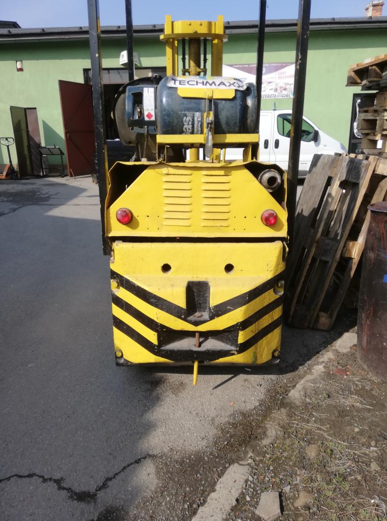 Żółty wózek widłowy Techmax z czarnymi pasami ostrzegawczymi, zaparkowany na zewnątrz na asfaltowym placu, widok z tyłu, obok palety i zielony budynek.