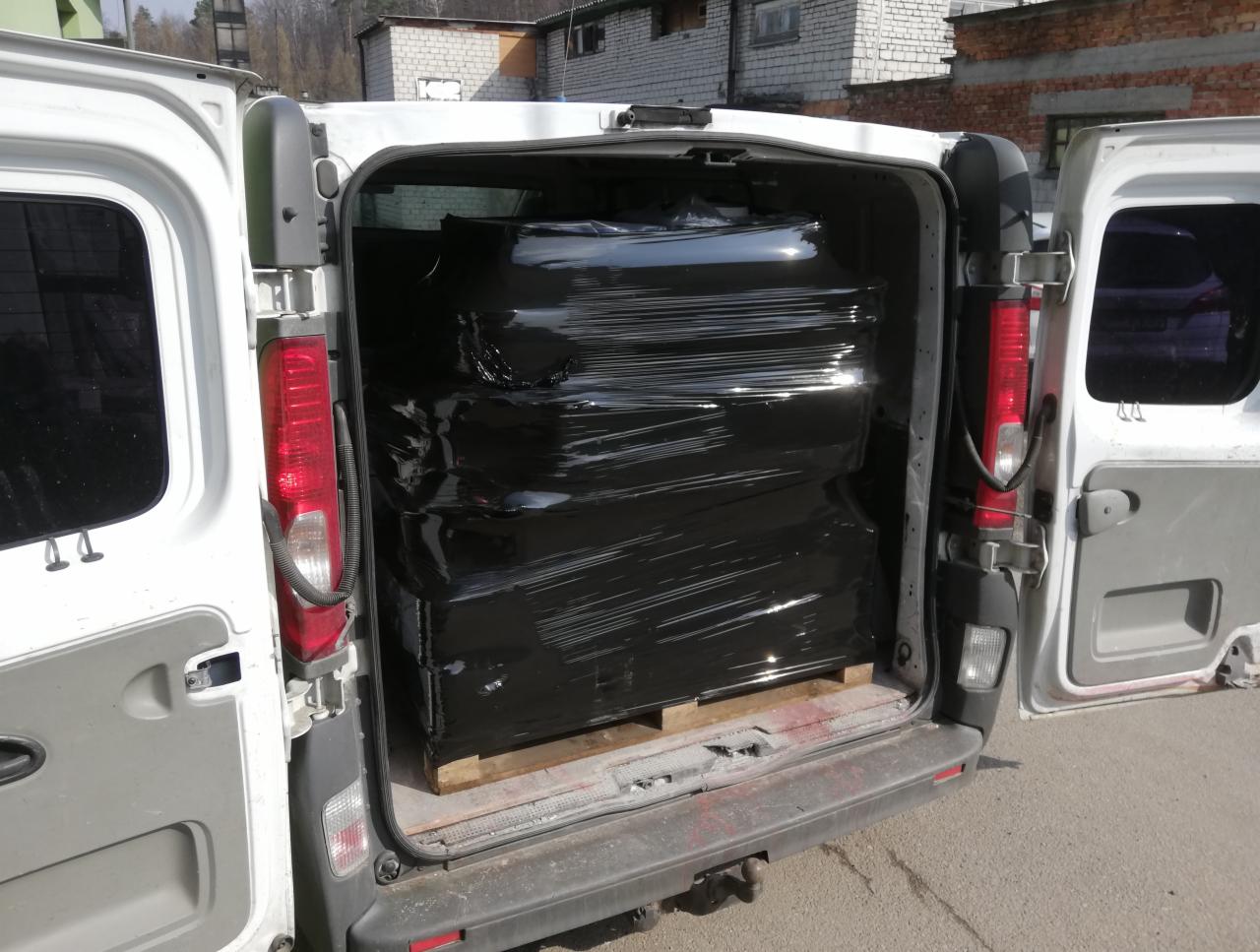 Wnętrze białego busa z otwartymi drzwiami, załadowanego czarnymi, owiniętymi folią pakunkami na drewnianej palecie.