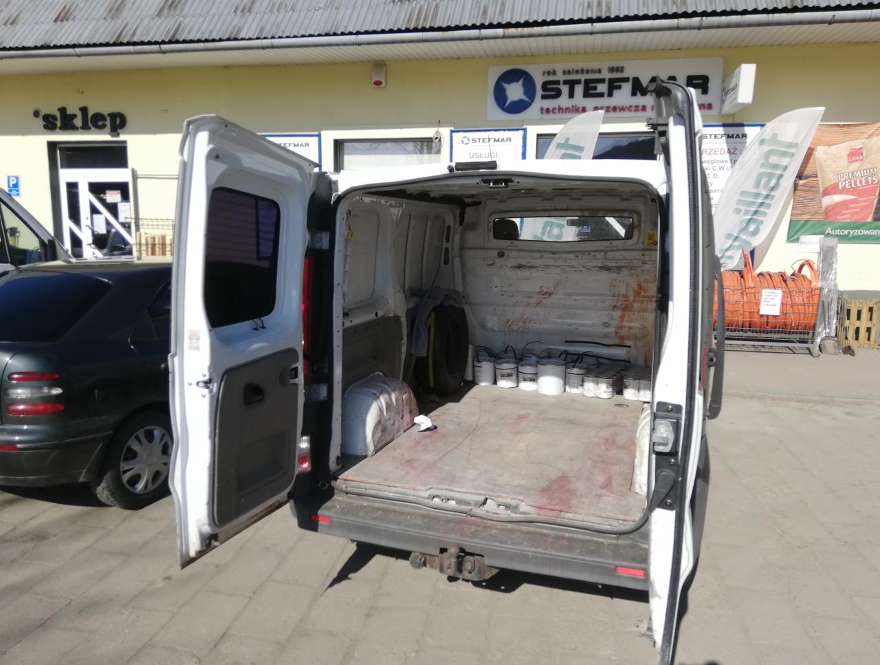 Otwarty bagażnik białego busa z widocznymi puszkami farb i zapasowym kołem, stojącego przed budynkiem z szyldem 'STEFMAR'.