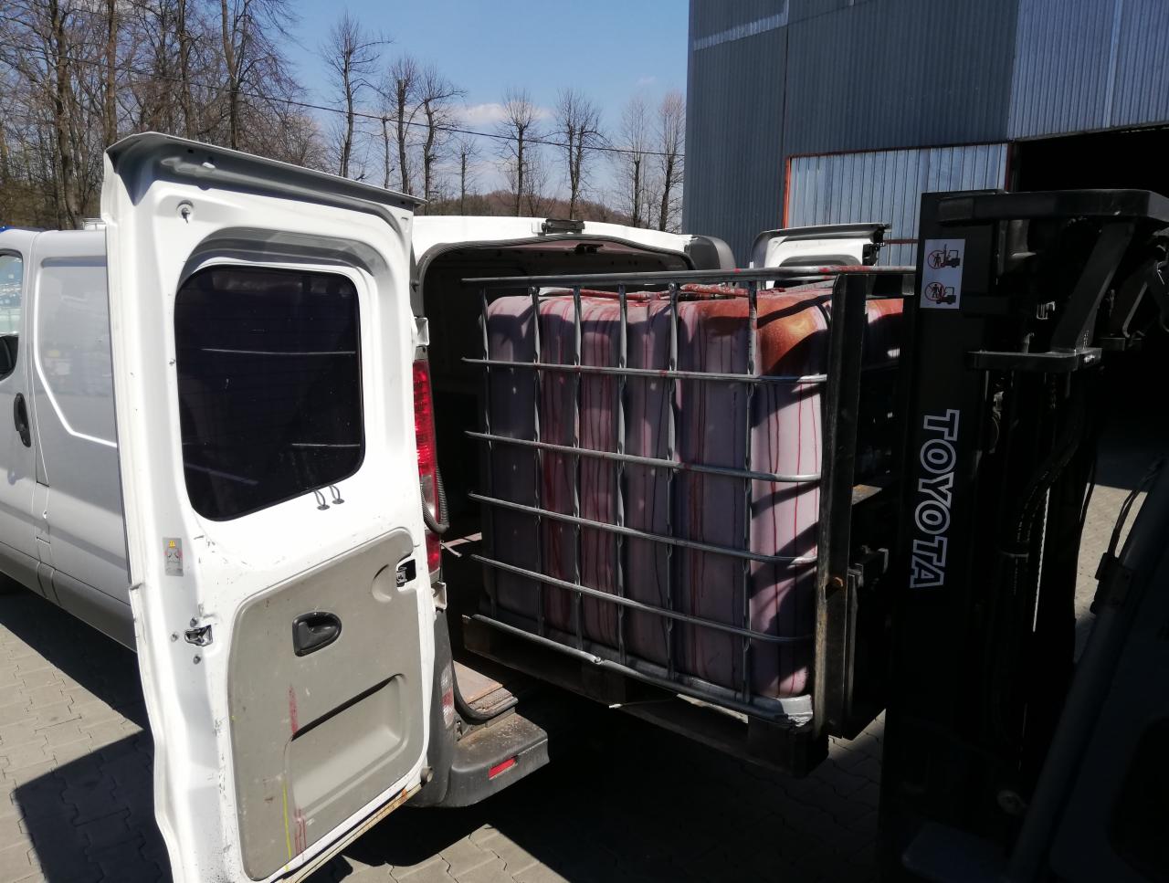 Wózek widłowy Toyota załadowuje kontener IBC z czerwonawą cieczą do białego busa z otwartymi drzwiami, widoczne drzewa w tle.