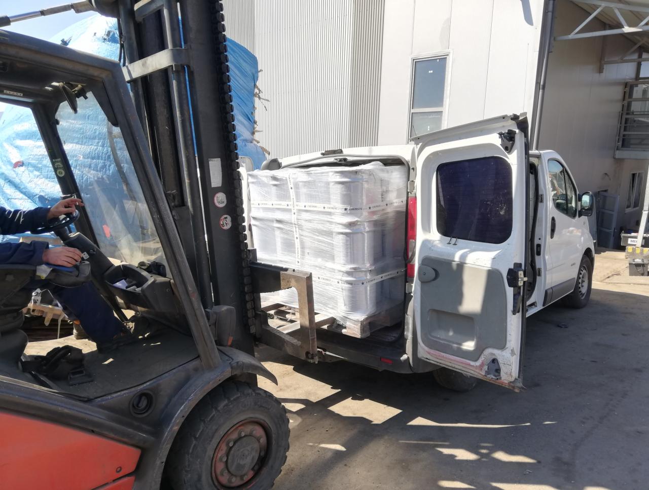Wózek widłowy ładuje paletę z zapakowanymi elementami do białego busa z otwartymi drzwiami, na tle blaszanej hali.