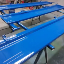 Dwie metalowe konstrukcje podnośników samochodowych, świeżo pomalowane na jaskrawy niebieski kolor, ustawione na kozłach, widoczne refleksy światła na gładkiej powierzchni farby.