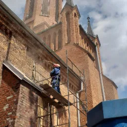 Pracownik w kombinezonie ochronnym i kasku na rusztowaniu czyści elewację ceglanego kościoła za pomocą strumienia pod ciśnieniem, prawdopodobnie przygotowując ją do malowania.