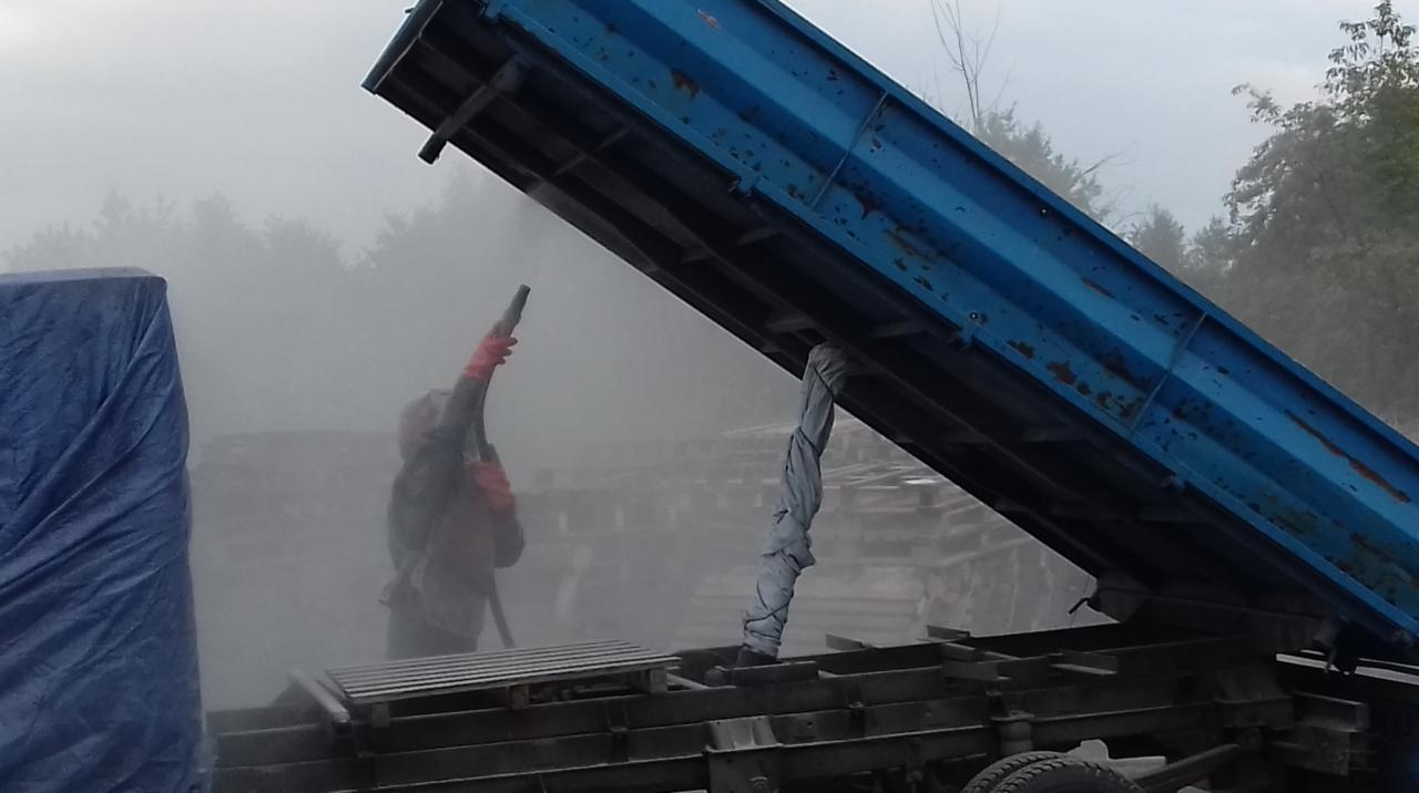 Pracownik w ochronnym kombinezonie i rękawicach, za pomocą pistoletu pneumatycznego, oczyszcza konstrukcję skrzyni wywrotki, z której unosi się obłok pyłu.