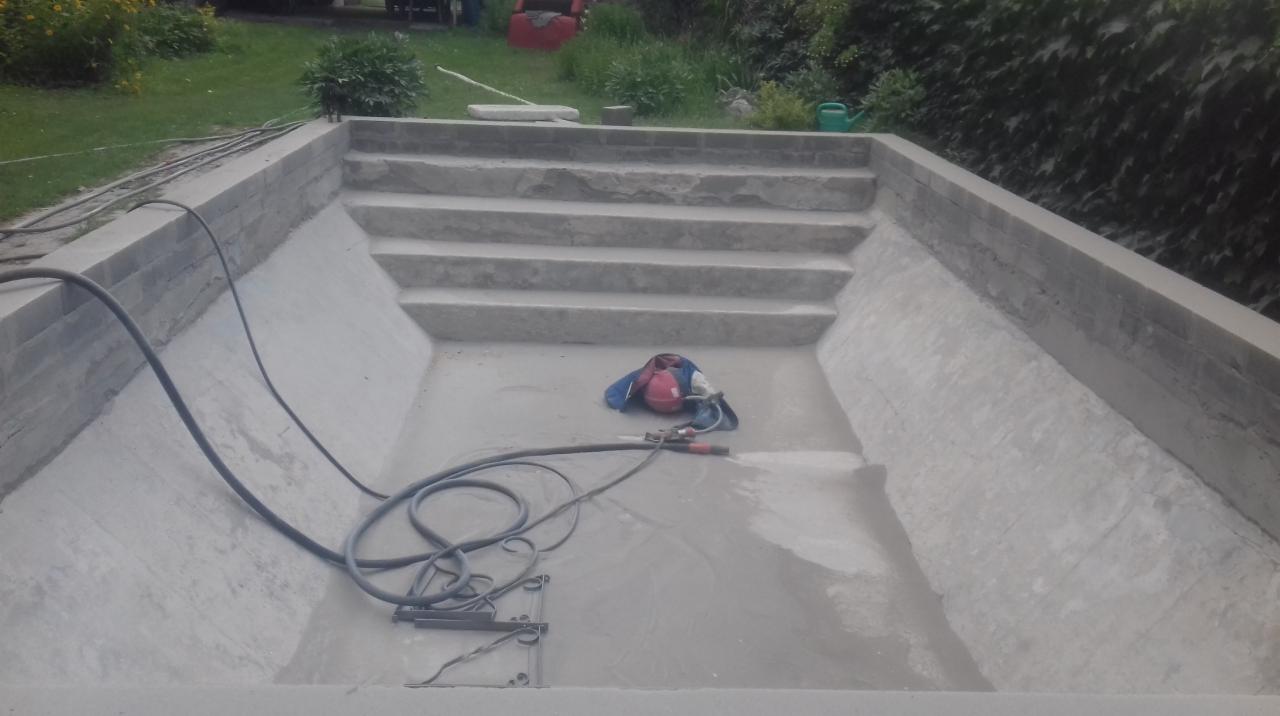 Betonowy basen w trakcie renowacji, widoczne schody, narzędzia i węże, prawdopodobnie przed czyszczeniem lub na etapie przygotowania powierzchni.