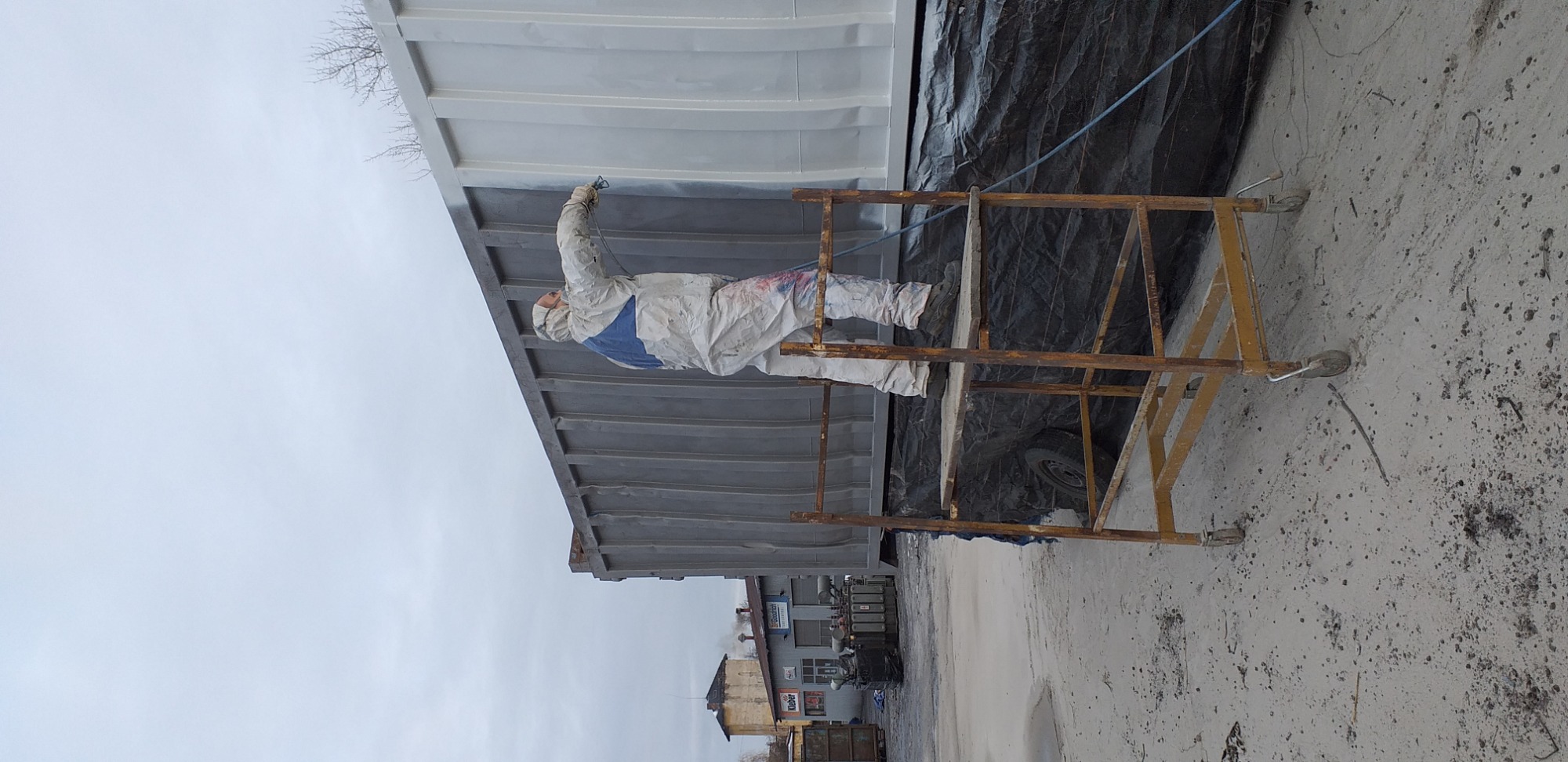 Pracownik w kombinezonie ochronnym, stojący na rusztowaniu, natryskowo nanosi farbę na kontener. Część kontenera jest już pomalowana na szaro.