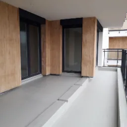 Nowoczesny balkon z szarą, gładką posadzką, drewnopodobnymi panelami ściennymi i czarną balustradą, widok z góry.