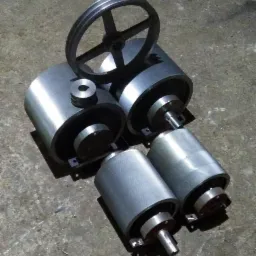 Srebrne, cylindryczne elementy metalowe, prawdopodobnie rolki i koło pasowe, ułożone na szorstkiej, betonowej powierzchni w warsztacie, z widocznymi śladami obróbki.