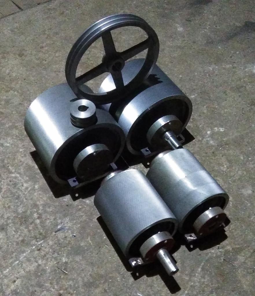 Srebrne, cylindryczne elementy metalowe, prawdopodobnie rolki i koło pasowe, ułożone na szorstkiej, betonowej powierzchni w warsztacie, z widocznymi śladami obróbki.