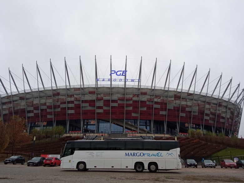 Biały autokar firmy Margo Travel zaparkowany przed Stadionem Narodowym w Warszawie, w tle pochmurne niebo.
