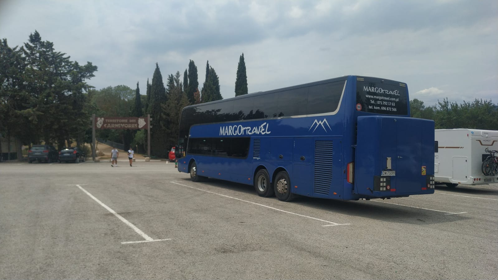 Niebieski, dwupokładowy autokar turystyczny Margo Travel zaparkowany na parkingu z widokiem na park Territoire des Carnivores w tle.