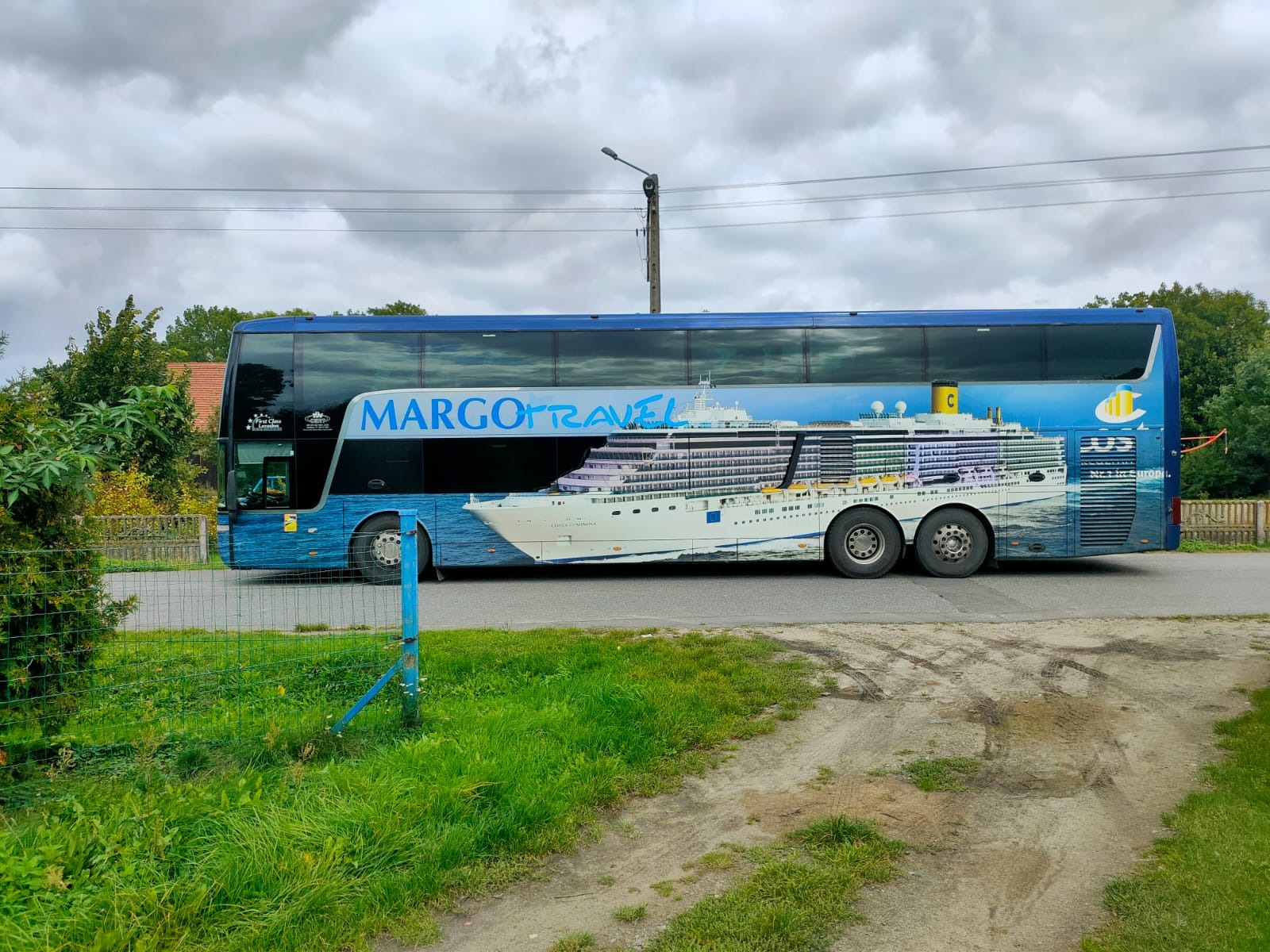 Dwupoziomowy autobus turystyczny z grafiką przedstawiającą statek wycieczkowy, zaparkowany przy drodze w pochmurny dzień.