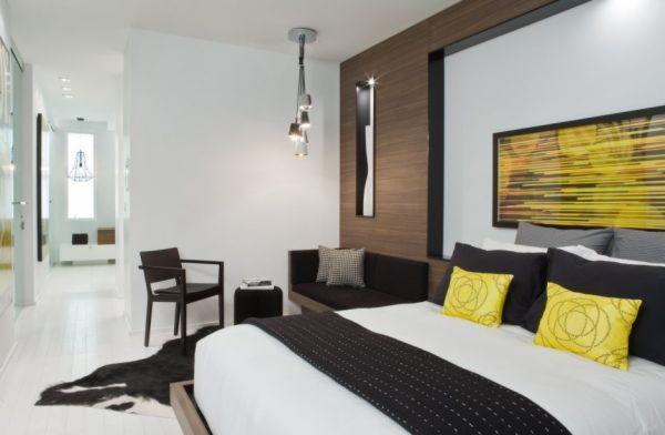 Nowoczesna sypialnia z białymi ścianami, drewnianą ścianą akcentującą, łóżkiem z czarno-białą narzutą i żółtymi poduszkami oraz czarnym krzesłem i dywanem imitującym skórę krowy.