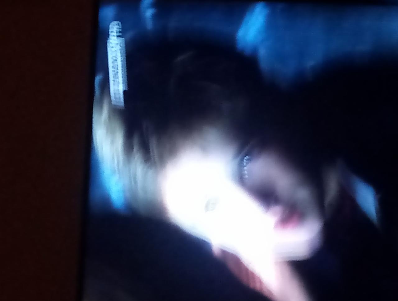 Rozmazane zdjęcie osoby z jasnymi włosami, zrobione prawdopodobnie z ekranu telewizora, z widocznymi artefaktami i słabym oświetleniem.