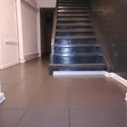 Klatka schodowa z ciemnymi schodami i szarą, wykafelkowaną podłogą. Widoczne białe rury na ścianie i wejście do innego pomieszczenia w tle.