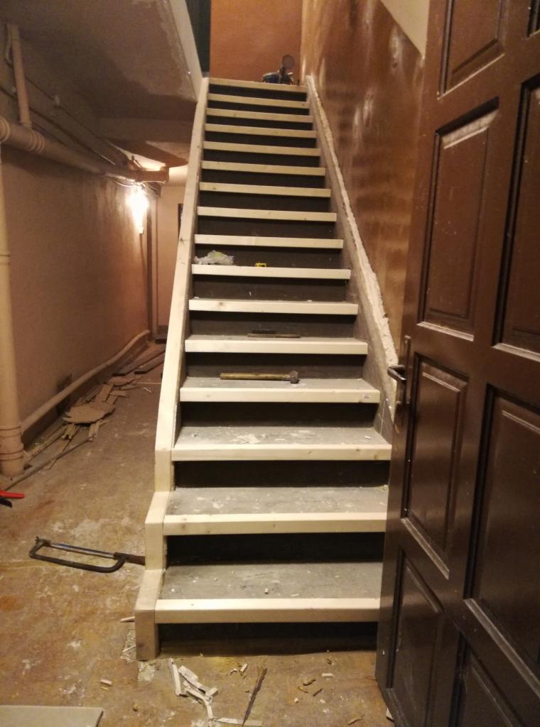 Wnętrze klatki schodowej w trakcie remontu: surowe drewniane schody z widocznymi śladami prac, młotek na jednym ze stopni, oraz częściowo otwarte ciemne drzwi po prawej stronie.