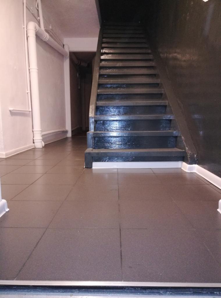 Klatka schodowa z ciemnymi schodami i szarą, wykafelkowaną podłogą. Widoczne białe rury na ścianie i wejście do innego pomieszczenia w tle.