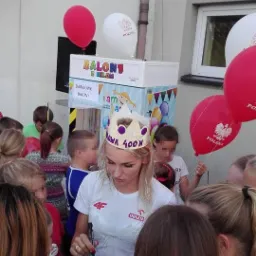 Kobieta w koronie rozdaje autografy dzieciom na tle dystrybutora balonów z napisem 'Darmowe balony' i logo Polski na balonach.