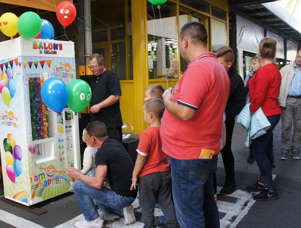 Automat z balonami na hel z motywem tęczy i napisem 'I am happy' obsługiwany przez mężczyznę w czarnej koszulce, w otoczeniu dzieci i dorosłych.