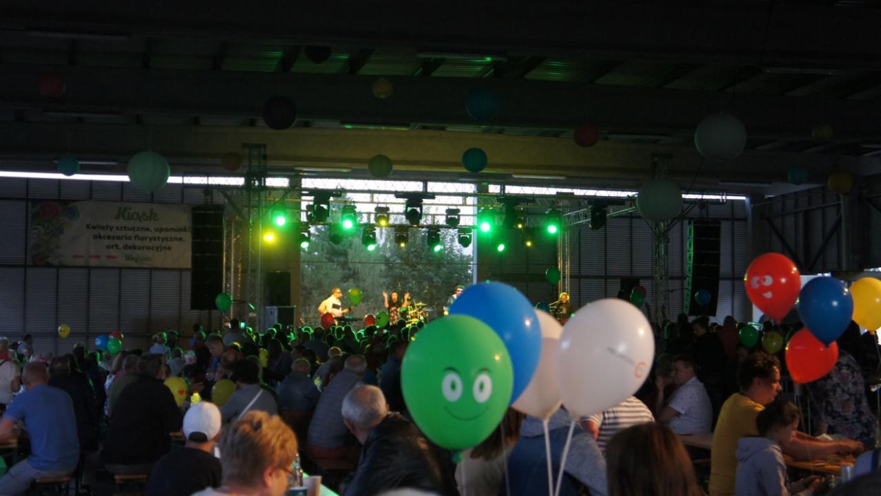 Koncert plenerowy z zespołem na scenie, oświetleniem i publicznością siedzącą przy stołach, dekorowany balonami, w tle baner reklamowy.