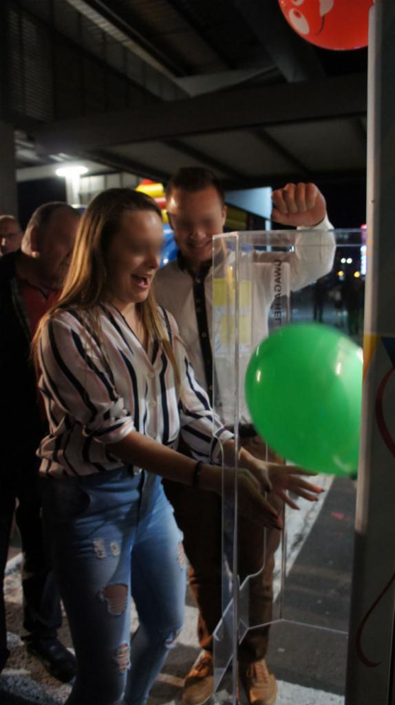 Uśmiechnięta kobieta w koszuli w paski i podartych jeansach bierze udział w zabawie z przezroczystym pudełkiem i balonami, mężczyzna w białej koszuli i brązowych spodniach podnosi rękę w geście...