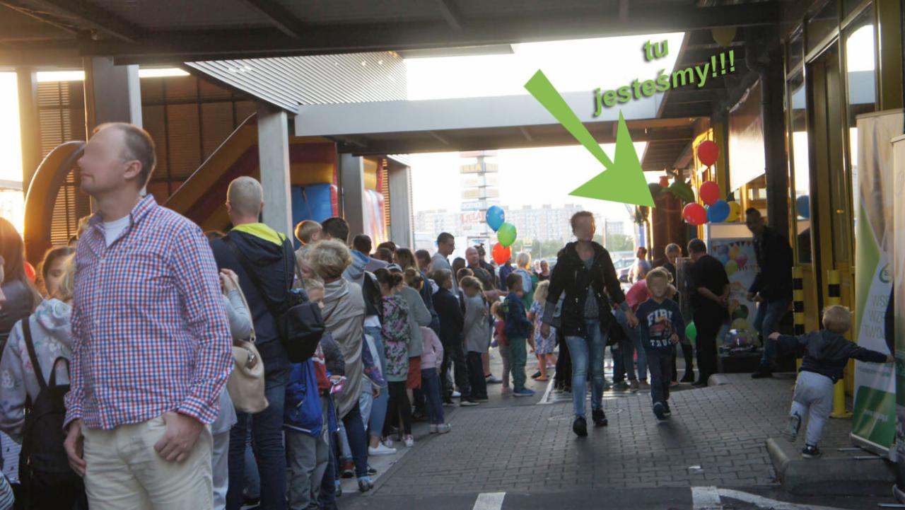 Tłum ludzi, głównie dzieci, zgromadzony przed wejściem, z balonami i dmuchańcem w tle, wskazany zieloną strzałką z napisem 'tu jesteśmy!!!'.
