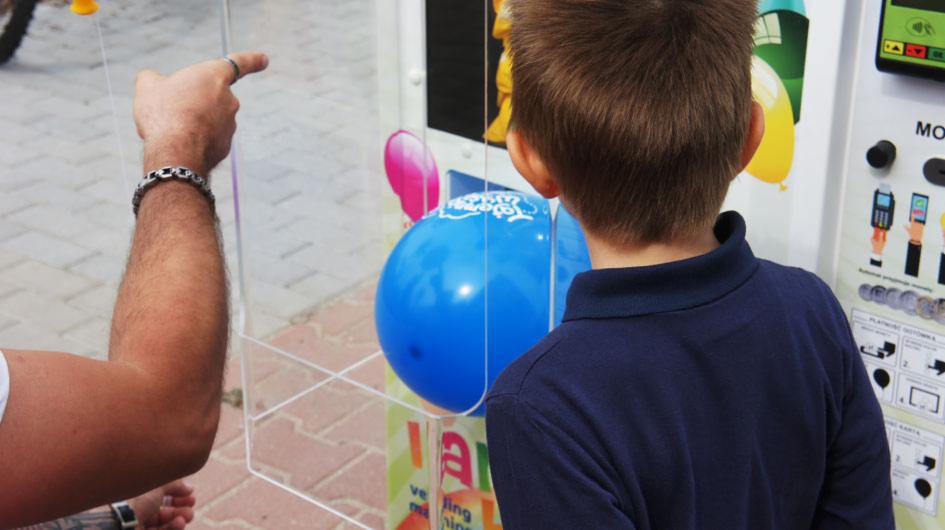 Chłopiec obserwuje niebieski balon w przezroczystej obudowie automatu zręcznościowego, wskazany palcem przez dorosłego.