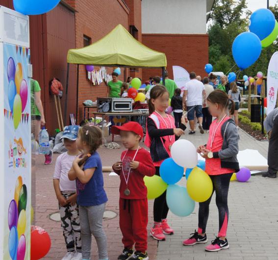 Grupa dzieci z medalami na szyjach i balonami w rękach stoi przed kolorową ścianką z grafiką balonów, w tle widoczny namiot, budynek i inni ludzie.