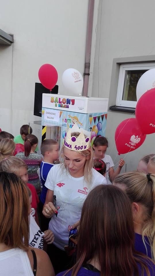 Kobieta w koronie rozdaje autografy dzieciom na tle dystrybutora balonów z napisem 'Darmowe balony' i logo Polski na balonach.