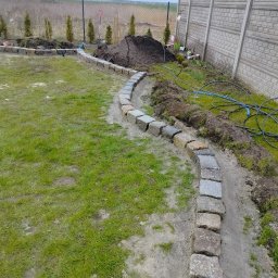Concrete Road Dominik Ochman - Wykopany rów z ułożoną kostką granitową wzdłuż trawnika, widoczne nasadzenia roślin, ziemia i ogrodzenie panelowe. Prace brukarskie w toku.