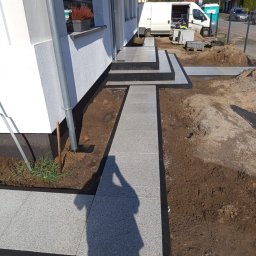 Concrete Road Dominik Ochman - Ścieżka z jasnoszarej kostki brukowej z czarnym obramowaniem prowadząca do wejścia domu. Widoczne schody z kostki i fragment terenu przed budynkiem w słoneczny dzień.