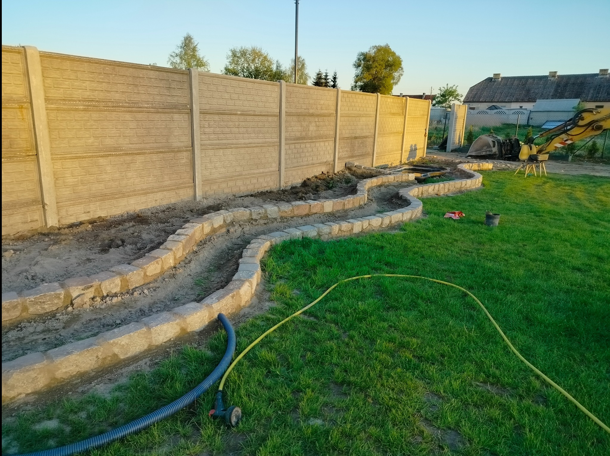 Betonowy płot z falującą kamienną obudową rabat. W tle widoczny sprzęt budowlany i dom. Żółty wąż leży na trawie. Ujęcie z dołu.