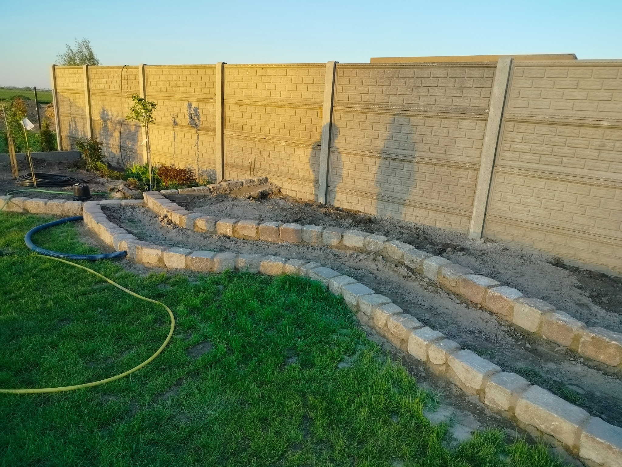 Ogród w trakcie aranżacji: betonowe ogrodzenie panelowe, obrzeża z kostki granitowej, przygotowane rabaty, węże ogrodowe. Widok z góry, wczesny wieczór.