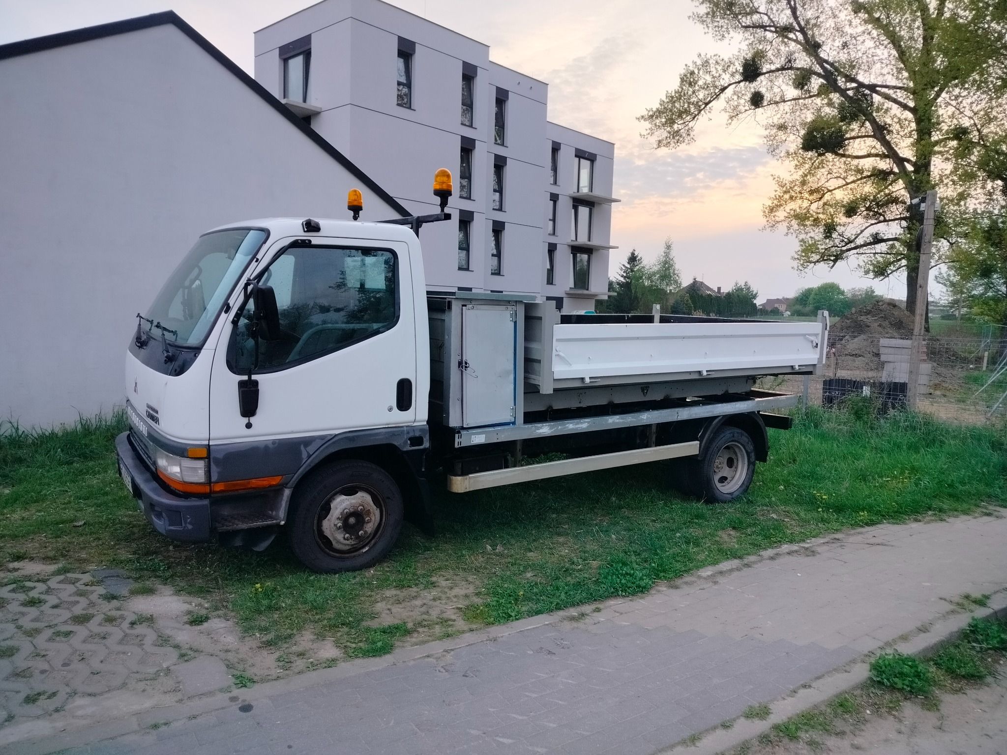 Biała ciężarówka z otwartą skrzynią ładunkową stoi na trawie przed nowoczesnym budynkiem mieszkalnym i drzewem, prawdopodobnie przygotowana do prac ziemnych.