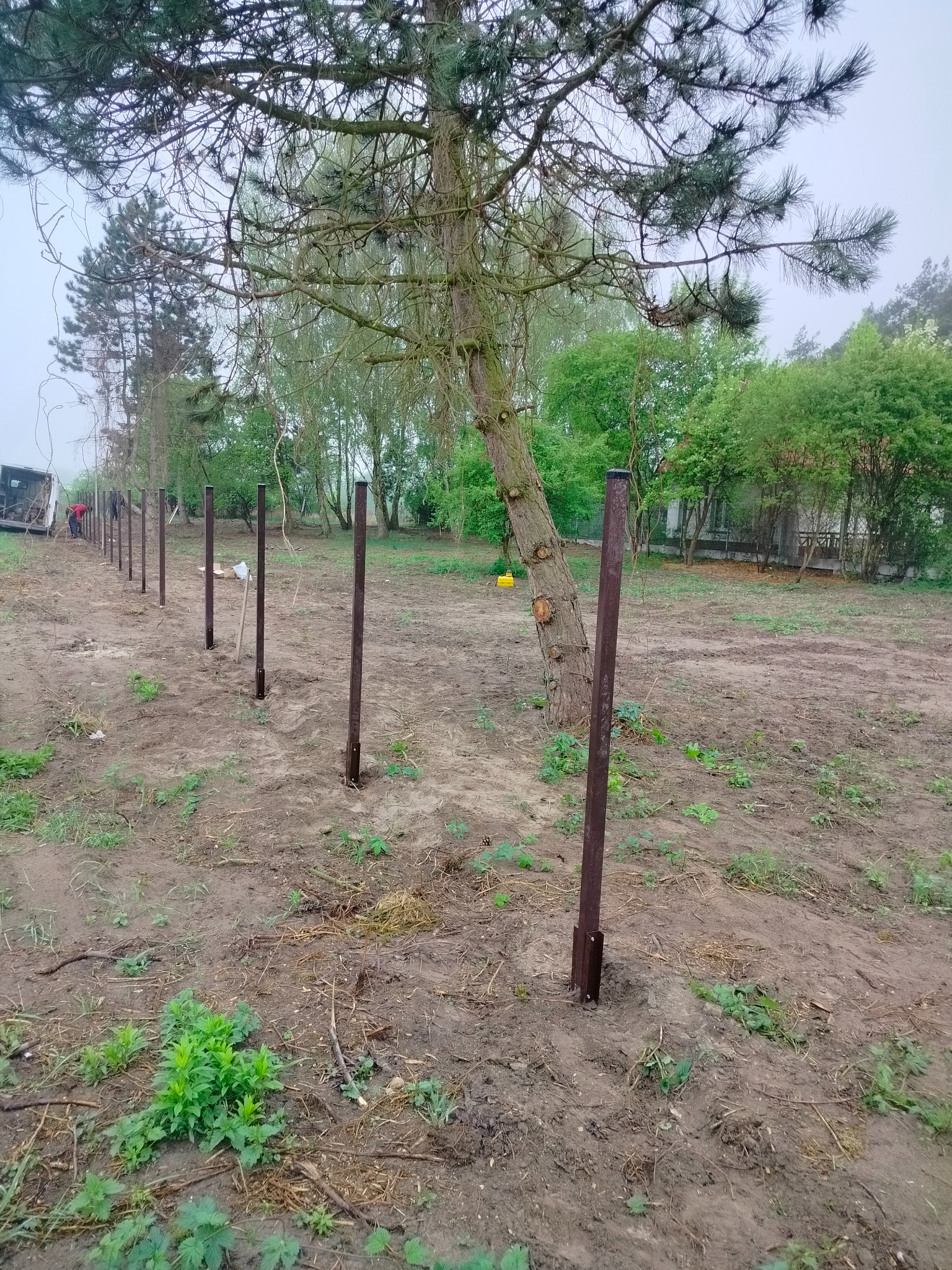 Montaż ogrodzenia: Metalowe słupki wkopane w ziemię, ustawione w rzędzie, tworzące linię przyszłego ogrodzenia na działce z drzewami w tle. Widoczny samochód dostawczy.