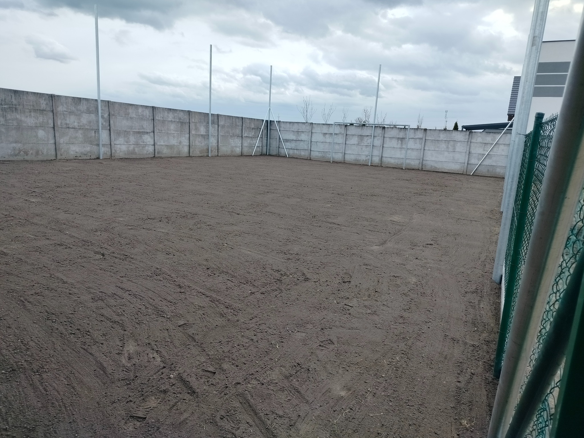 Teren wyrównany ziemią, otoczony betonowym murem i metalowymi słupkami. Przygotowanie pod budowę lub ogród. Pochmurne niebo.