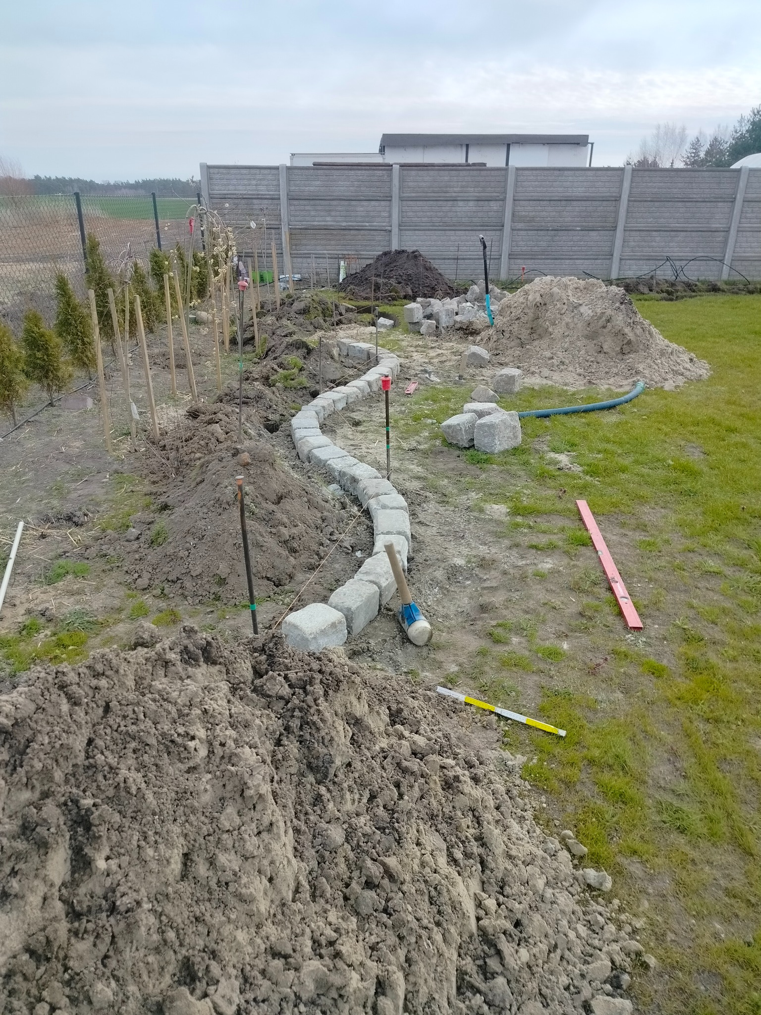 Krawężnik z kostki granitowej w trakcie budowy, wyznaczający ścieżkę w ogrodzie. Widoczne narzędzia i materiały budowlane, świeżo posadzone tuje. Tło: betonowe ogrodzenie.