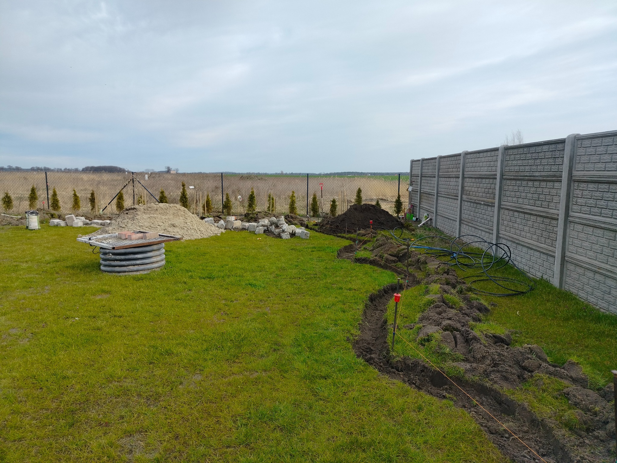 Przygotowanie do drenażu na trawniku: wykopany rów, studzienka betonowa, kostka brukowa i ogrodzenie panelowe w tle. Widoczne węże i narzędzia.