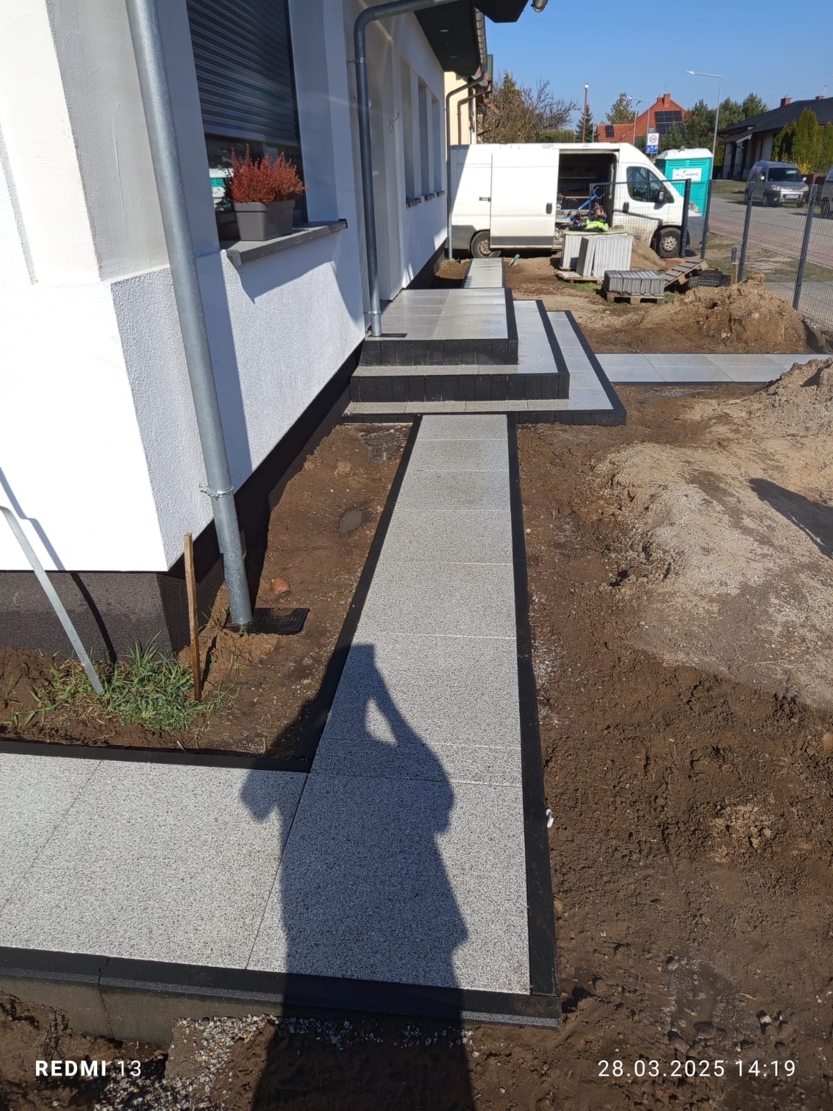 Ścieżka z jasnoszarej kostki brukowej z czarnym obramowaniem prowadząca do wejścia domu. Widoczne schody z kostki i fragment terenu przed budynkiem w słoneczny dzień.