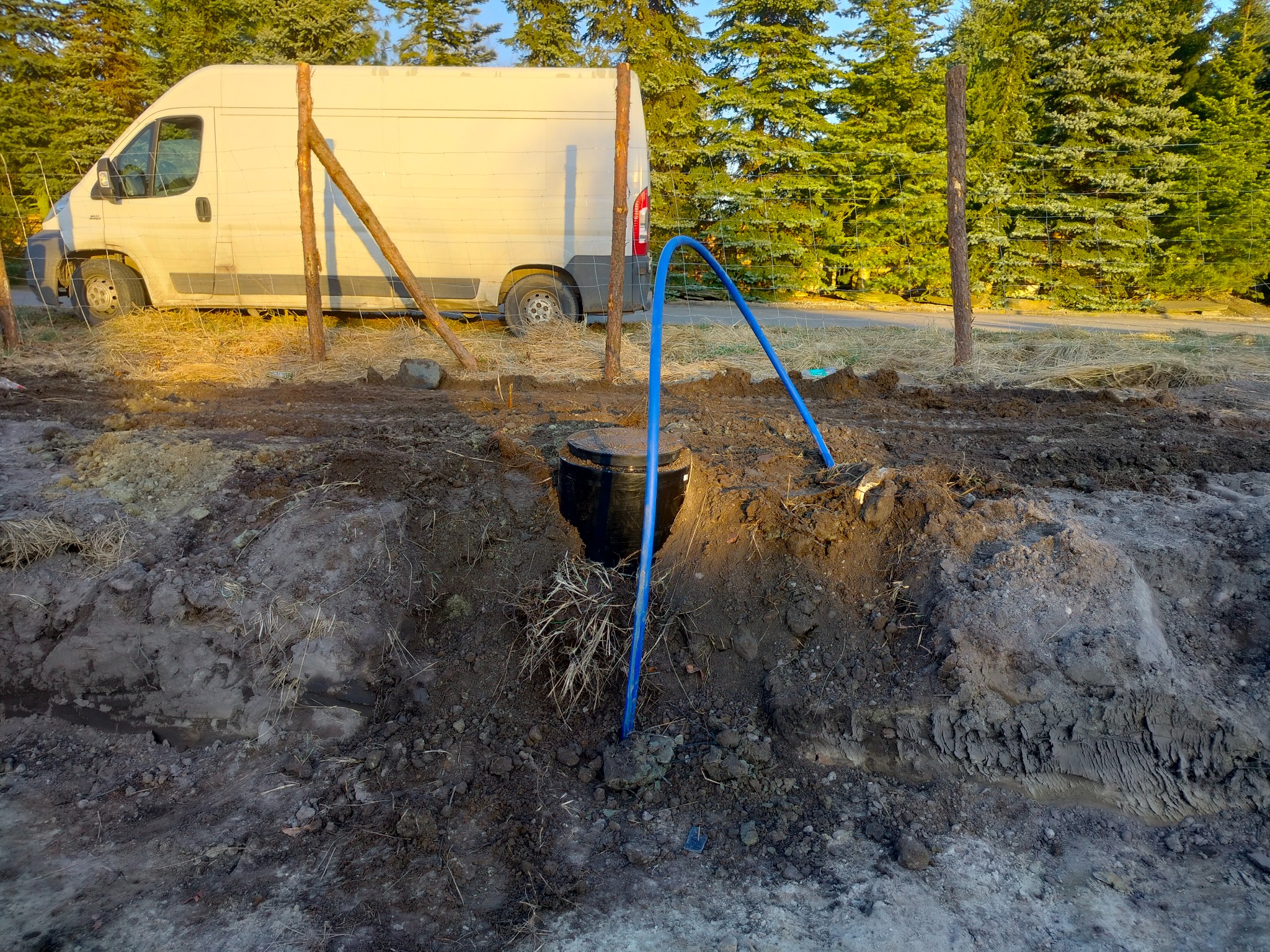 Studzienka kanalizacyjna z niebieską rurą w wykopie, na tle białego busa i ogrodzenia z drewnianych słupków. Widoczne drzewa iglaste.