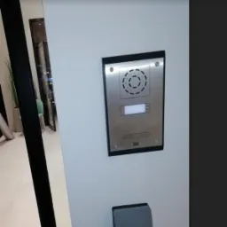 Nowoczesny panel domofonu ze stali nierdzewnej, zamontowany na białej ścianie, z widocznym przyciskiem dzwonka i mikrofonem, w tle częściowo widoczne wnętrze budynku.