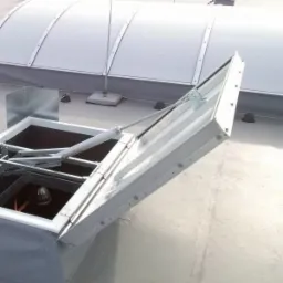 Otwarty, biały świetlik dachowy z siłownikiem hydraulicznym na płaskim dachu, widoczny fragment dachu i kopuła świetlika w tle.