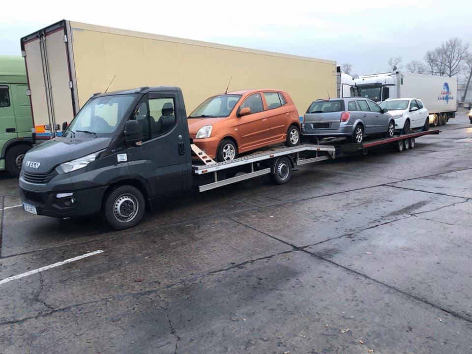 Ciemnoszara laweta Iveco przewozi trzy samochody osobowe: pomarańczowy hatchback, szary kombi i biały hatchback. Na drugim planie widoczne zaparkowane ciężarówki z naczepami.