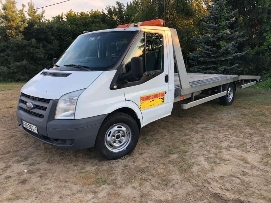 Biały Ford Transit przystosowany jako laweta, z pomarańczowym światłem ostrzegawczym na dachu, zaparkowany na piaszczystym terenie z widocznym napisem 'Pomoc Drogowa' na boku.