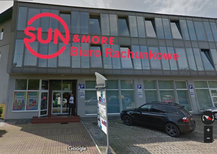 Dwupiętrowy budynek z szarą elewacją i dużymi oknami, z widocznym logo firmy 'SUN & MORE' oraz napisem 'Biuro Rachunkowe'. Na parkingu przed budynkiem zaparkowany czarny samochód. Przed wejściem...