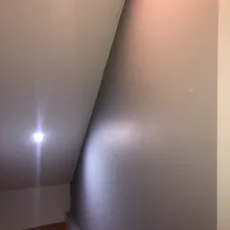 Wnętrze klatki schodowej po malowaniu, widoczna gładka szara ściana pod skosem, biały sufit z wbudowanym punktowym oświetleniem LED.